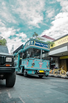 A bright blue tour bus labeled 'Bandung Tour on Bus' is parked on a cobblestone street. There is a yellow bicycle next to a shop with a sign reading 'Mardika.' The sky is partly cloudy, and another vehicle is partially visible in the foreground.