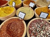 Various types of spices are displayed in wooden bowls, each labeled with their names. The spices include vibrant colors such as yellow, red, and mixtures of different spices. The labels identify them as Falafel Spice, Fish Spice, Harissa Spice, and Mixed Pepper, among others.
