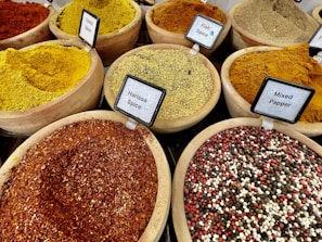 Various types of spices are displayed in wooden bowls, each labeled with their names. The spices include vibrant colors such as yellow, red, and mixtures of different spices. The labels identify them as Falafel Spice, Fish Spice, Harissa Spice, and Mixed Pepper, among others.