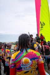 A group of people gather near the water, wearing colorful jerseys with vibrant patterns and logos. One large, brightly colored sail is visible in the background, indicating a sailing event or competition. The crowd appears diverse, with various hairstyles and clothing choices, suggesting a lively and festive atmosphere.