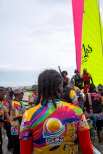 A group of people gather near the water, wearing colorful jerseys with vibrant patterns and logos. One large, brightly colored sail is visible in the background, indicating a sailing event or competition. The crowd appears diverse, with various hairstyles and clothing choices, suggesting a lively and festive atmosphere.