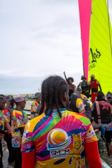 A group of people gather near the water, wearing colorful jerseys with vibrant patterns and logos. One large, brightly colored sail is visible in the background, indicating a sailing event or competition. The crowd appears diverse, with various hairstyles and clothing choices, suggesting a lively and festive atmosphere.