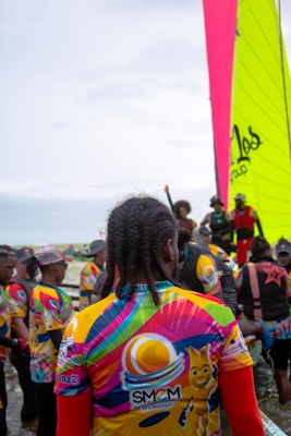A group of people gather near the water, wearing colorful jerseys with vibrant patterns and logos. One large, brightly colored sail is visible in the background, indicating a sailing event or competition. The crowd appears diverse, with various hairstyles and clothing choices, suggesting a lively and festive atmosphere.