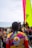 A group of people gather near the water, wearing colorful jerseys with vibrant patterns and logos. One large, brightly colored sail is visible in the background, indicating a sailing event or competition. The crowd appears diverse, with various hairstyles and clothing choices, suggesting a lively and festive atmosphere.