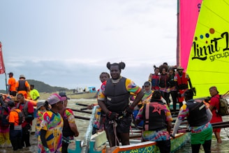A group of people dressed in colorful sports attire gathers around a large canoe with eye-catching multicolored sails, preparing for or concluding a boat race on a cloudy day near a shoreline.
