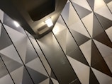 The image features a modern interior with a geometric pattern of triangular shapes on the wall. The surfaces have a metallic appearance, with a gradient of shades from light to dark. Two ceiling lights are visible, casting soft illumination across the surfaces.