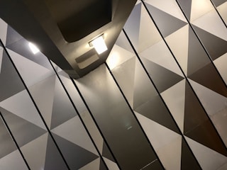 The image features a modern interior with a geometric pattern of triangular shapes on the wall. The surfaces have a metallic appearance, with a gradient of shades from light to dark. Two ceiling lights are visible, casting soft illumination across the surfaces.