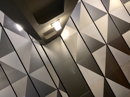 The image features a modern interior with a geometric pattern of triangular shapes on the wall. The surfaces have a metallic appearance, with a gradient of shades from light to dark. Two ceiling lights are visible, casting soft illumination across the surfaces.