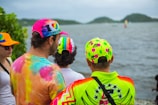A group of people stand by a waterfront, wearing colorful, patterned clothing and hats. The hats are adorned with various bright designs and messages. The background includes a cloudy sky and distant hills across the water.