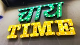 A brightly lit sign with two parts, featuring the text in both a local language and English. The upper part of the sign is in green with illuminated bulbs, while the lower part spells 'TIME' in yellow with similar bulb lighting.