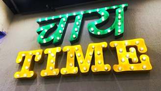 A brightly lit sign with two parts, featuring the text in both a local language and English. The upper part of the sign is in green with illuminated bulbs, while the lower part spells 'TIME' in yellow with similar bulb lighting.