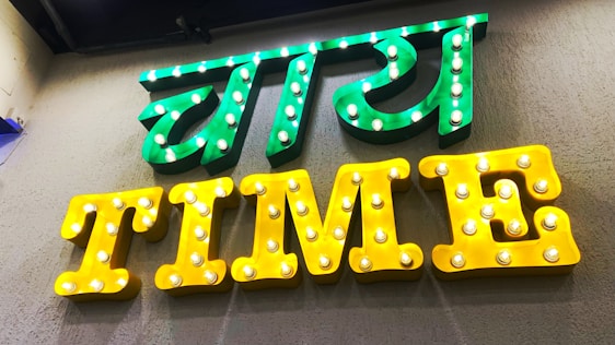 A brightly lit sign with two parts, featuring the text in both a local language and English. The upper part of the sign is in green with illuminated bulbs, while the lower part spells 'TIME' in yellow with similar bulb lighting.