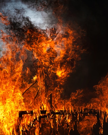 Flames engulf a structure, creating a dramatic and intense scene. The bright orange and yellow colors of the fire contrast sharply against the dark background, with smoke swirling upward. A distinctive detail, such as a hand-like shape, emerges amid the fiery chaos.