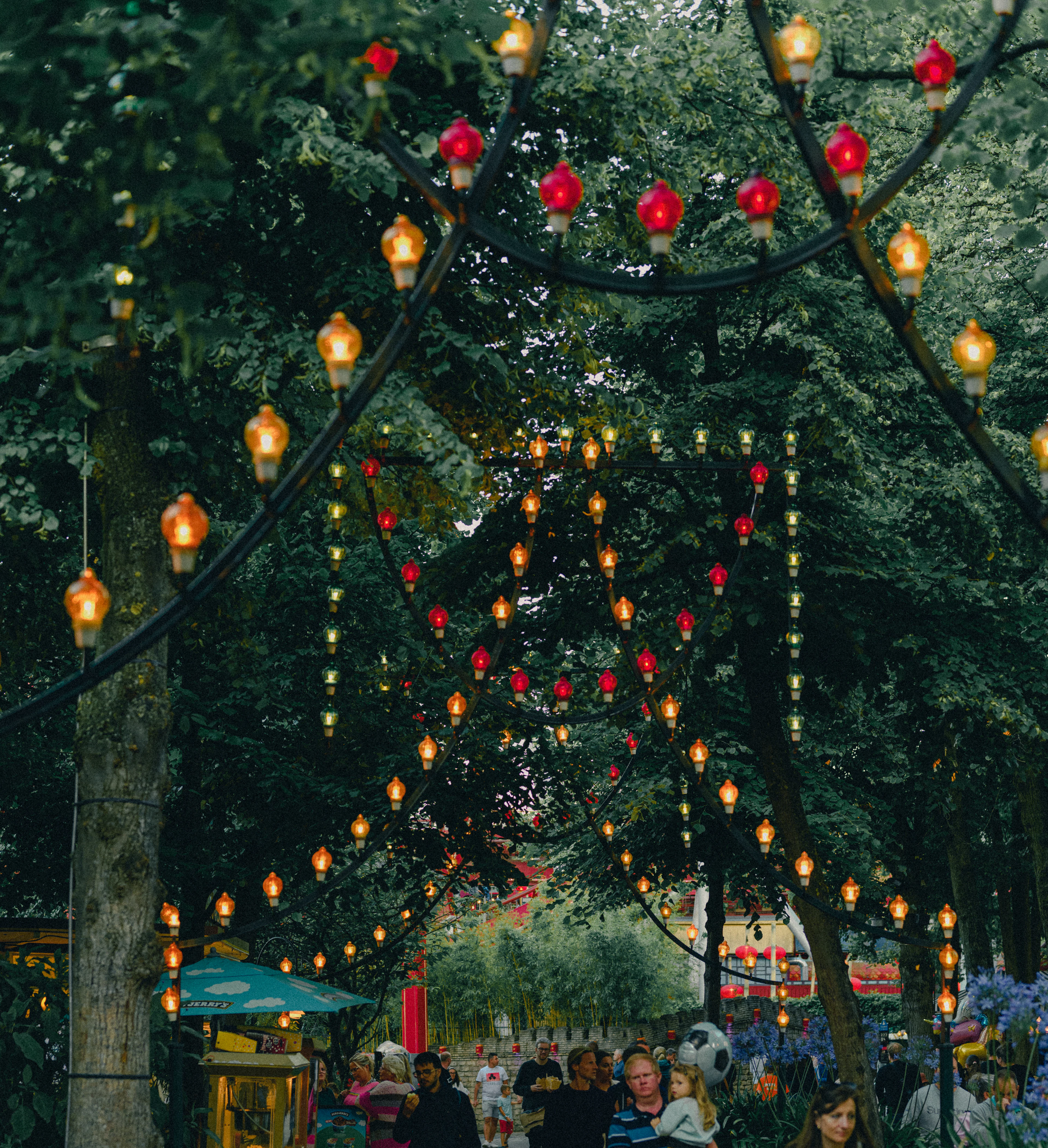 Strings of red and amber lanterns hung in long arcs through the summer trees of Tivoli Gardens in Copenhagen, glowing against dark green leaves as visitors walk the path below