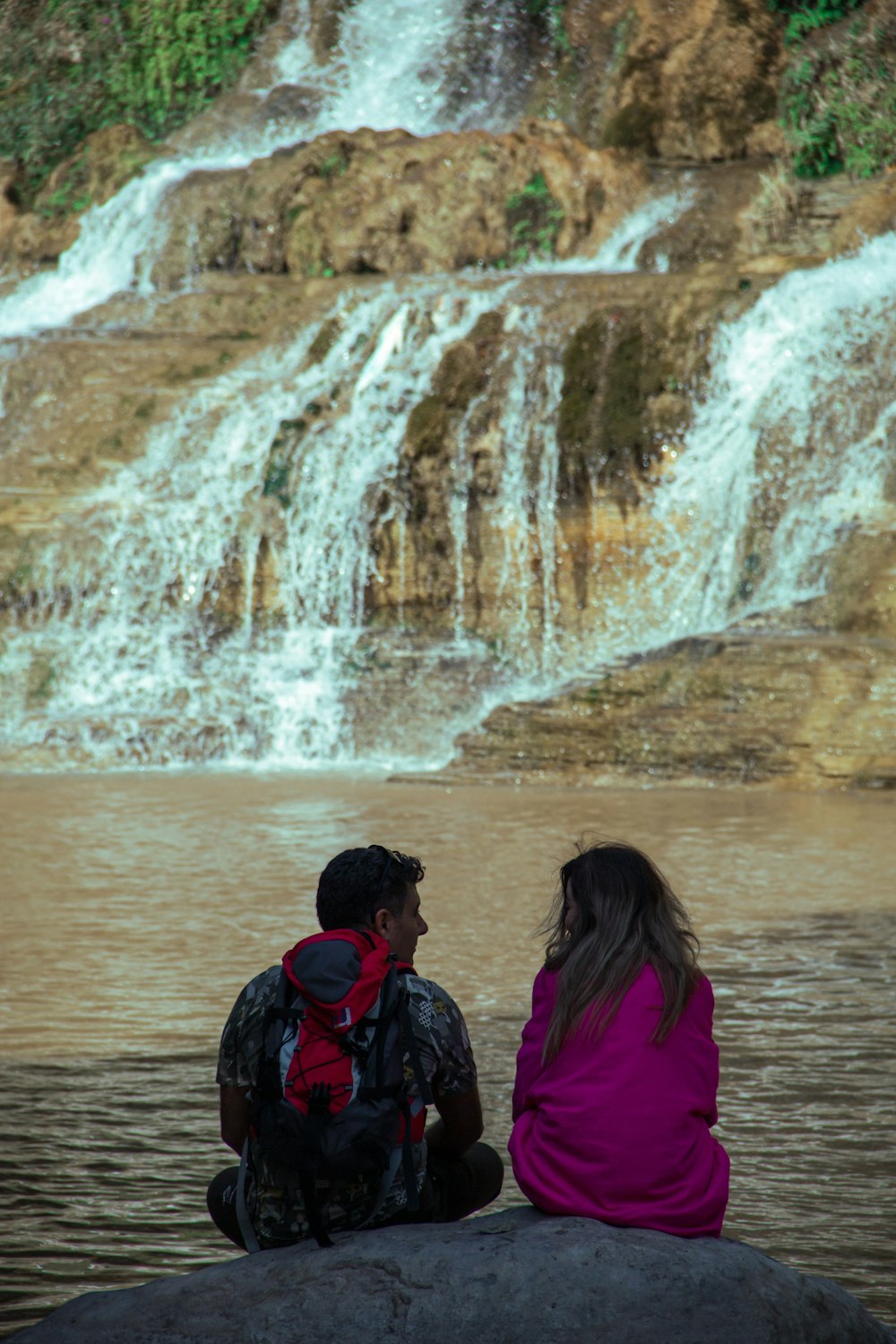 a man and woman sitting on a rock looking at a waterfall
