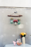 A rustic wall decoration with three glass jars hanging from a wooden stick, each containing different types of flowers. Below, a table covered with a light cloth displays a tall vase with yellow and orange flowers, alongside a glass container filled with colorful candies and a stack of pink napkins.