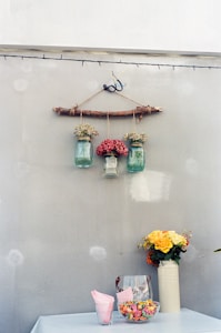 A rustic wall decoration with three glass jars hanging from a wooden stick, each containing different types of flowers. Below, a table covered with a light cloth displays a tall vase with yellow and orange flowers, alongside a glass container filled with colorful candies and a stack of pink napkins.
