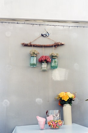 A rustic wall decoration with three glass jars hanging from a wooden stick, each containing different types of flowers. Below, a table covered with a light cloth displays a tall vase with yellow and orange flowers, alongside a glass container filled with colorful candies and a stack of pink napkins.