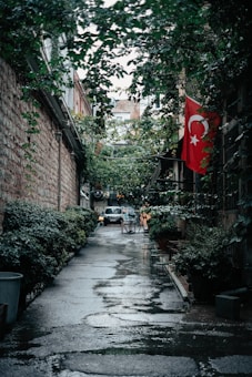 An alleyway lined with leafy green plants, and a Turkish flag displayed on the right side. The ground is wet, suggesting recent rain, and a parked car is visible in the distance. Chairs and tables are set up along the right side, hinting at an outdoor seating area, perhaps for a café.