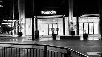 A modern building with a glass facade illuminated from the inside, showcasing a sign that reads 'Foundry' above the entrance. The area is largely empty, with a few potted plants and architectural elements visible. The lighting suggests it is nighttime.