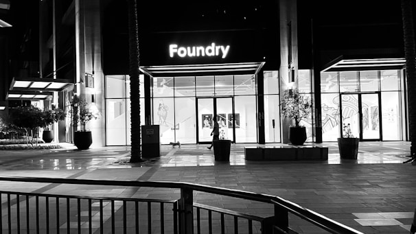 A modern building with a glass facade illuminated from the inside, showcasing a sign that reads 'Foundry' above the entrance. The area is largely empty, with a few potted plants and architectural elements visible. The lighting suggests it is nighttime.