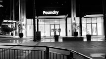 A modern building with a glass facade illuminated from the inside, showcasing a sign that reads 'Foundry' above the entrance. The area is largely empty, with a few potted plants and architectural elements visible. The lighting suggests it is nighttime.
