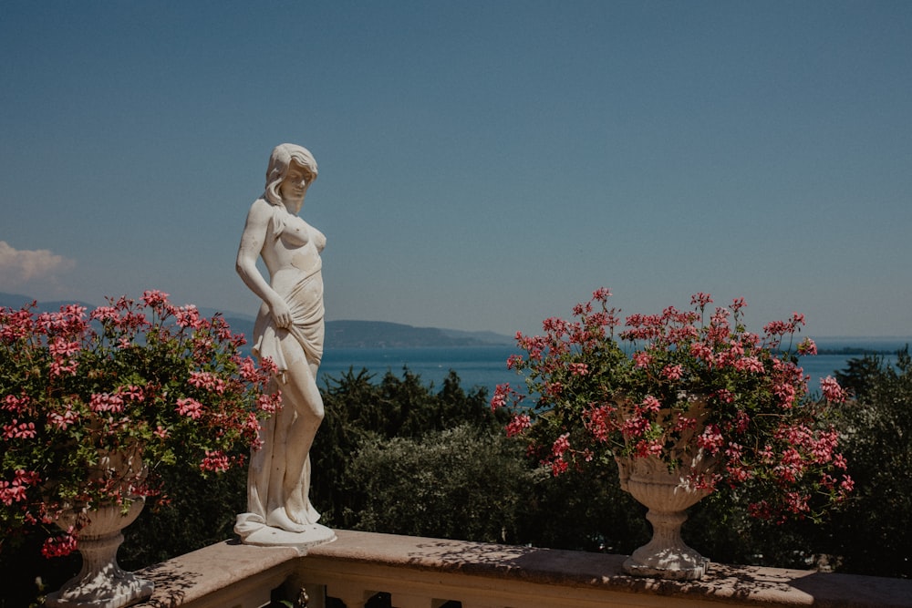 a statue of a person on a ledge with flowers and a body of water in the background