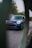 A blue Mini Cooper parked on a street, partially obscured by green foliage in the foreground. The car has distinctive circular headlights and a sporty front grille, with slightly overcast weather and trees in the background.