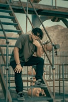 A person is sitting thoughtfully on a set of industrial metal stairs. The background includes industrial equipment and a fence, with a muted, earthy color palette. The individual is dressed casually in dark pants and a dark shirt, with sunglasses, creating a reflective pose.