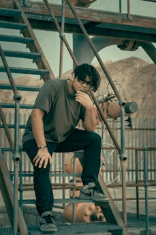 A person is sitting thoughtfully on a set of industrial metal stairs. The background includes industrial equipment and a fence, with a muted, earthy color palette. The individual is dressed casually in dark pants and a dark shirt, with sunglasses, creating a reflective pose.