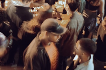 A bustling crowd with people engaged in conversation and movement. The scene is slightly blurred, creating a sense of motion and energy. Individuals appear to be socializing in a lively environment.