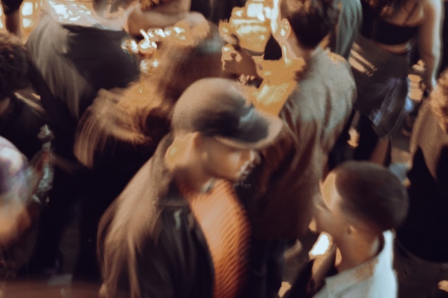 A bustling crowd with people engaged in conversation and movement. The scene is slightly blurred, creating a sense of motion and energy. Individuals appear to be socializing in a lively environment.