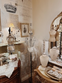 An elegant boutique display featuring vintage-inspired jewelry and decor. Mannequins are adorned with necklaces and tiaras, sitting on a richly decorated wooden dresser. The backdrop includes ornate wallpaper with classical scripts and crown illustrations, and a framed historical portrait. A mirror reflects part of the setup, enhancing the luxurious ambiance.