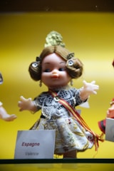 A doll dressed in traditional attire, featuring a lace-covered dress with intricate patterns and a red and yellow sash. The doll has styled brown hair and a flower accessory on top. It is placed against a bright yellow background, possibly in a display setting. A label with the words 'Espagne' and 'Valence' is visible next to it, suggesting a Spanish theme.
