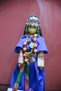 A decorative doll dressed in traditional attire with intricate accessories. The doll features a long, vibrant blue robe with a colorful underlayer and patterned sleeves. It is adorned with various beads and coins, forming an elaborate necklace and headpiece, creating a rich and cultural appearance.