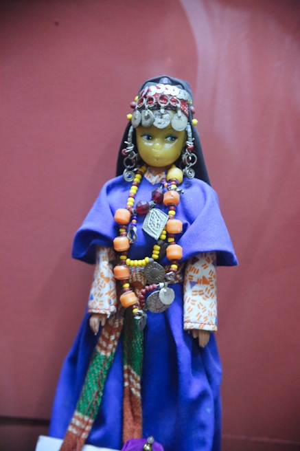 A decorative doll dressed in traditional attire with intricate accessories. The doll features a long, vibrant blue robe with a colorful underlayer and patterned sleeves. It is adorned with various beads and coins, forming an elaborate necklace and headpiece, creating a rich and cultural appearance.