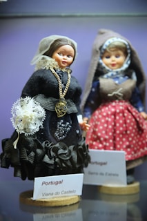 Two dolls dressed in traditional Portuguese costumes are displayed. The doll in the foreground wears a black outfit with intricate lace and bead detailing, accessorized with a gold chain. Its head is covered with a sheer lace veil. The second doll in the background is dressed in a blue and red polka dot outfit with a brown vest and a headscarf featuring a geometric pattern.