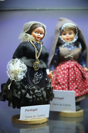 Two dolls dressed in traditional Portuguese costumes are displayed. The doll in the foreground wears a black outfit with intricate lace and bead detailing, accessorized with a gold chain. Its head is covered with a sheer lace veil. The second doll in the background is dressed in a blue and red polka dot outfit with a brown vest and a headscarf featuring a geometric pattern.