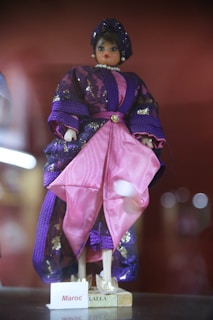 A doll dressed in traditional attire featuring a combination of purple and pink fabrics adorned with shiny accents. The doll is wearing a headpiece and a pearl necklace, with a nameplate at the base labeled 'Maroc Lalla'.