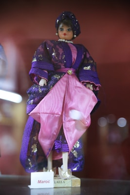 A doll dressed in traditional attire featuring a combination of purple and pink fabrics adorned with shiny accents. The doll is wearing a headpiece and a pearl necklace, with a nameplate at the base labeled 'Maroc Lalla'.