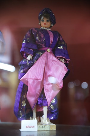 A doll dressed in traditional attire featuring a combination of purple and pink fabrics adorned with shiny accents. The doll is wearing a headpiece and a pearl necklace, with a nameplate at the base labeled 'Maroc Lalla'.
