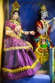 Two intricately designed dolls dressed in vibrant traditional attire, adorned with ornate jewelry and headpieces. One doll wears a purple dress with golden accents, while the other sports a multicolored outfit with similar decorative elements. Both figures are positioned in a setting that enhances their cultural aesthetic.