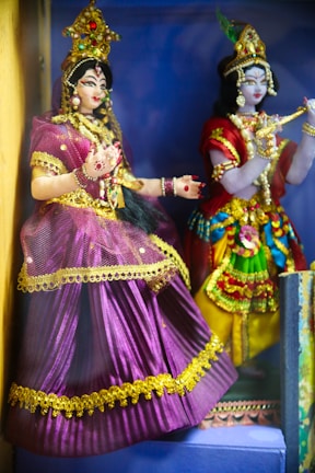 Two intricately designed dolls dressed in vibrant traditional attire, adorned with ornate jewelry and headpieces. One doll wears a purple dress with golden accents, while the other sports a multicolored outfit with similar decorative elements. Both figures are positioned in a setting that enhances their cultural aesthetic.