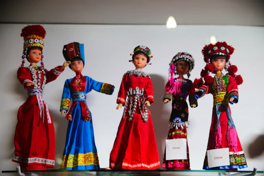 Five decorative dolls are dressed in traditional, colorful costumes showcasing intricate embroidery and accessories. Each doll's attire reflects a different style rich in cultural design, featuring vibrant colors and detailed ornamentation. The dolls are arranged in a display, emphasizing the artistic craftsmanship.