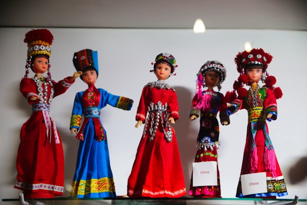 Five decorative dolls are dressed in traditional, colorful costumes showcasing intricate embroidery and accessories. Each doll's attire reflects a different style rich in cultural design, featuring vibrant colors and detailed ornamentation. The dolls are arranged in a display, emphasizing the artistic craftsmanship.