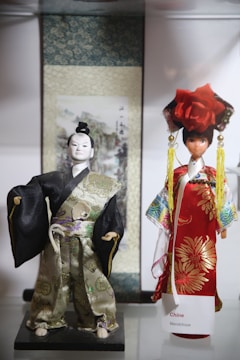 Two dolls dressed in traditional costumes are displayed. The doll on the left is wearing a black and gold ornate kimono-like outfit, with detailed embroidery and a tied belt. The doll on the right is clad in a vibrant red and gold dress adorned with a large flower headpiece and long tassels. They are positioned in front of a decorative scroll with an intricate backdrop.