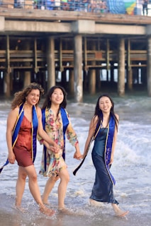 Three women are walking along the shoreline, holding hands and smiling, with a pier in the backdrop. They are dressed in various dresses and wearing graduation stoles.