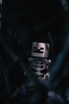 A toy skeleton figure with a square head, featuring pixelated features, is situated in a dark and mysterious setting. The surrounding environment is obscured and shadowy, adding an eerie and enigmatic atmosphere.