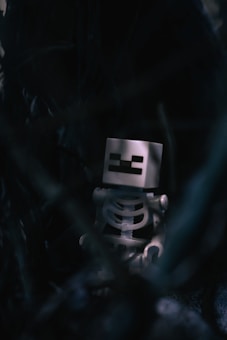 A toy skeleton figure with a square head, featuring pixelated features, is situated in a dark and mysterious setting. The surrounding environment is obscured and shadowy, adding an eerie and enigmatic atmosphere.