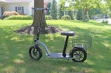An electric scooter with a black frame and a seat is parked on a grassy lawn. It features a wire basket attached to the rear. The background includes a tree, surrounding greenery, and a residential house partially visible with a cream facade and a window.